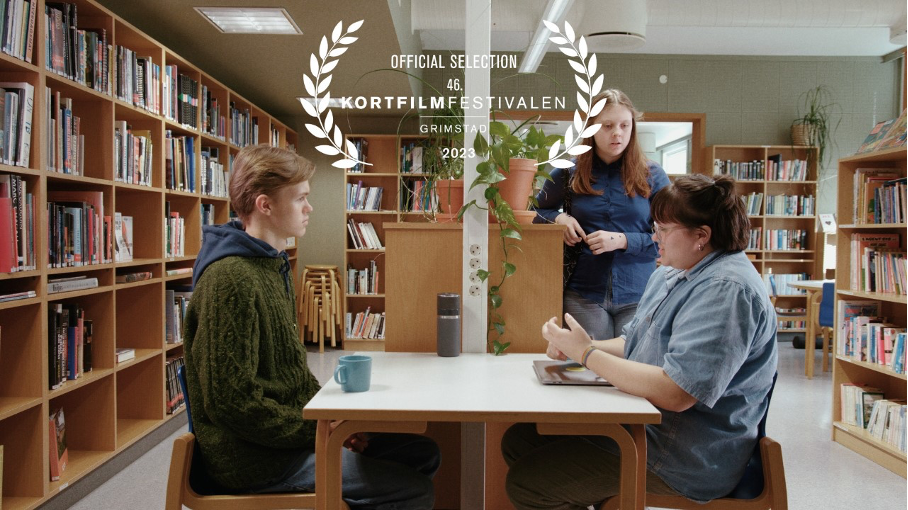 Bilde fra kortfilmen Shh som viser som elever som sitter ved et bord på biblioteket til Skeisvang vgs og en elev som står ved bordet - Klikk for stort bilde